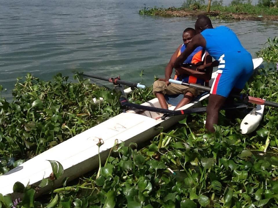 A coach helps a Para rowing athlete settle into an adapted boat on the water's edge.